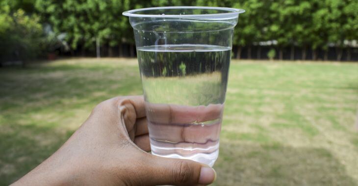 A person holding a glass of water over a lawn with dead grass.