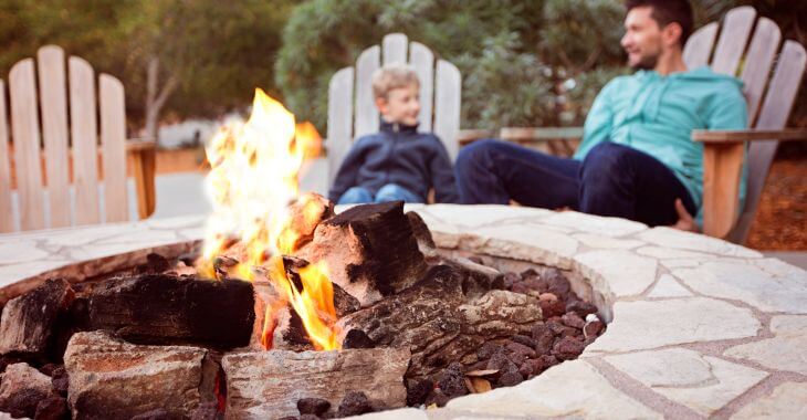 A father and a little boy sitting on the wooden armchairs by a firepit in the backyard and chatting